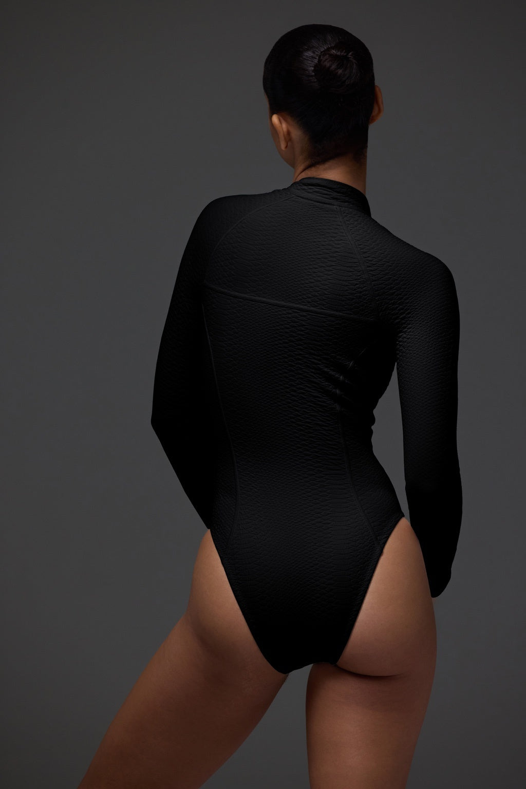 A person wearing a textured Sophie Surfsuit by Gigi C Bikinis stands with their back to the camera. The snakeskin-embossed fabric complements their bun hairstyle against a plain dark background.