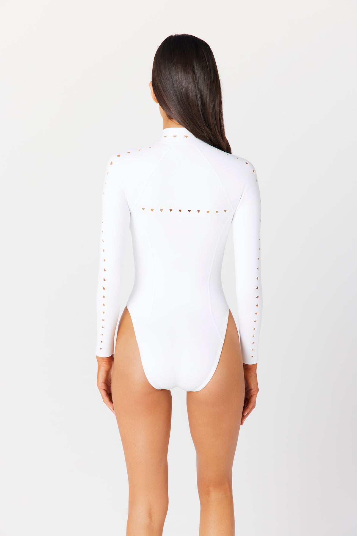 A person with long brown hair faces away, wearing a white Riley Surfsuit Full Coverage by Gigi C Bikinis. It features geometric cutout designs on the arms and shoulders, with a high-cut leg adding style against a plain white background.