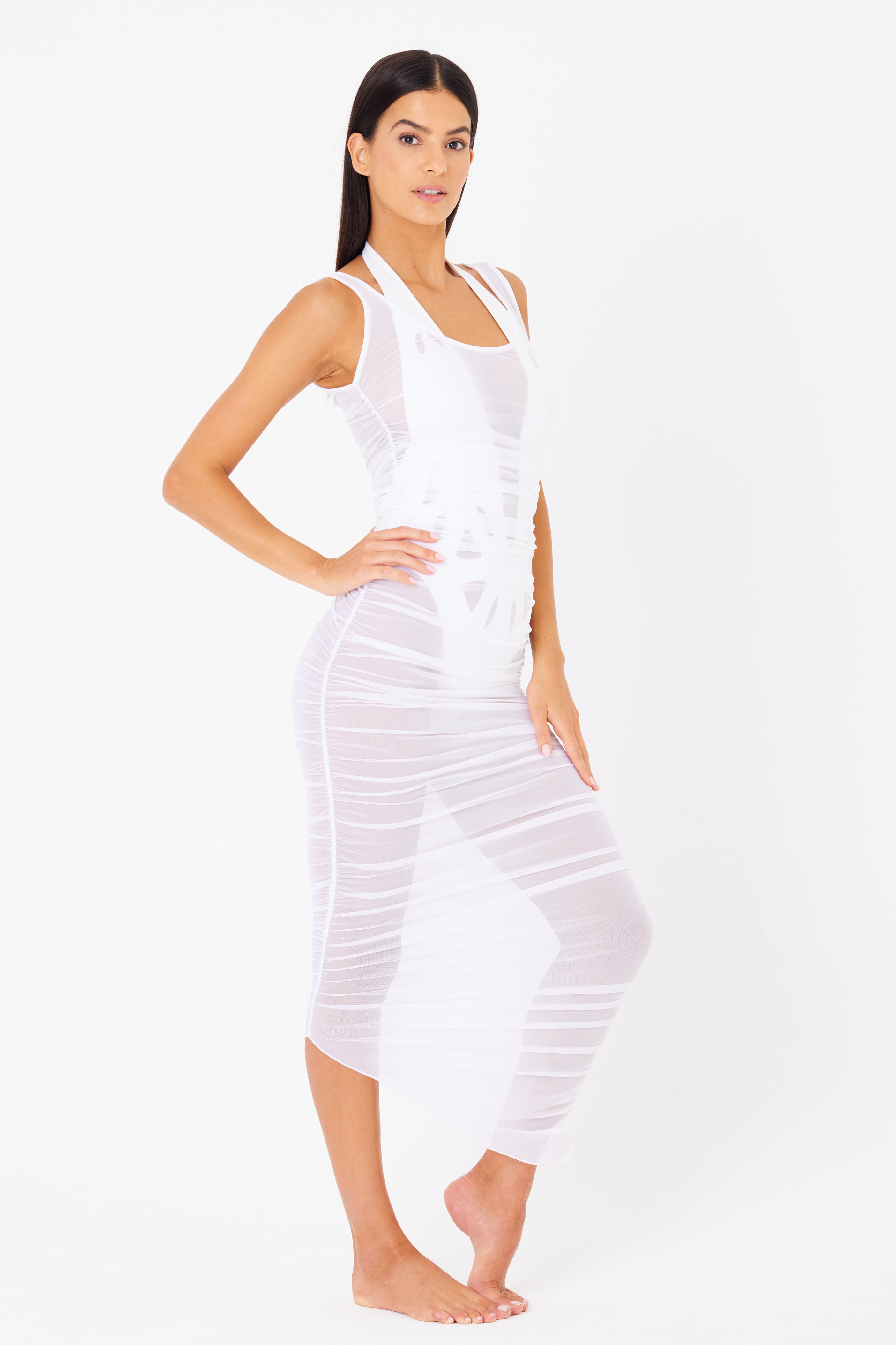 A person with long dark hair poses barefoot against a white backdrop, wearing the Stevie Midi Dress by Gigi C Bikinis. The sheer white dress features an intricate design and an asymmetrical hemline as they gaze at the camera with a relaxed demeanor.